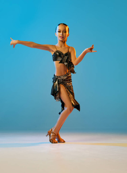 Samba dance. Beautiful little girl in stage attire dancing latino madern ballroom dance over blue background. Emotions, music, motion, education, professional dance school concept - Photo, image