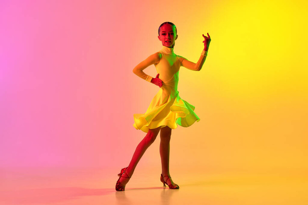 Music in movements. Emotional little girl in yellow stage dress dancing classical ballroom dance over gradient pink-yellow background in neon light filter. Concept of beauty, professional dances - Photo, Image