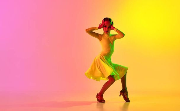 Music in movements. Emotional little girl in yellow stage dress dancing classical ballroom dance over gradient pink-yellow background in neon light filter. Concept of beauty, professional dances - Photo, image
