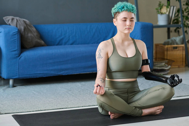 Junge Frau mit prothetischem Arm meditiert in Lotusposition mit geschlossenen Augen im Raum - Foto, Bild