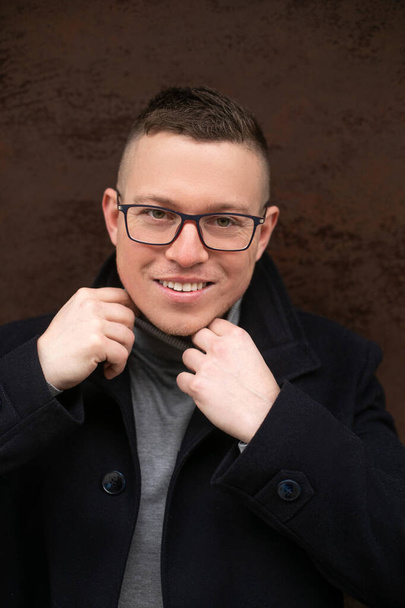 Handsome smiling man in glasses and black coat. Happy business man straighten turtleneck collar outdoors in cold day. Elegant middle aged person. Casual male outwear, stylish street style. - Photo, Image