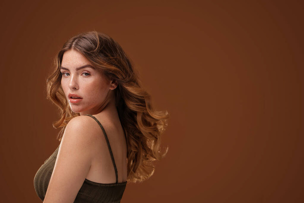 Beauty woman with long wavy hairstyle and natural freckles looking at the camera. Girl with delicate makeup. A lot of copy space on the brown studio background.  - Foto, imagen