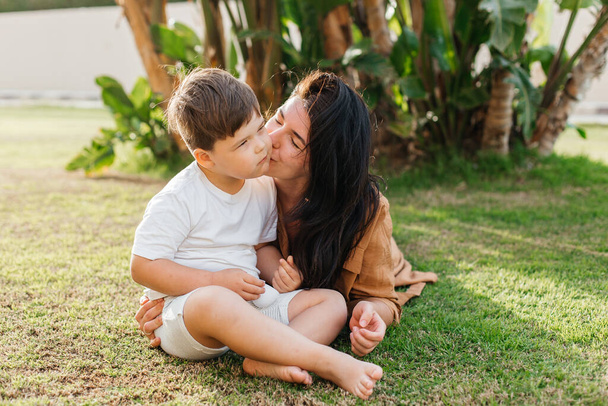 Happy smiing mother with son sitting on grass in park and smiling in camera. Parenting, family, children development, and fun outdoors in nature. High quality photo - Photo, image
