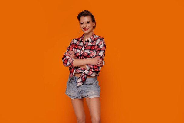 Beautiful young woman with red lipstick looking at the camera, posing in short jeans and fashionable shirt. Orange studio background with copy space.  - Foto, Bild