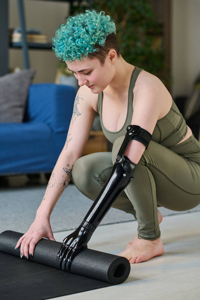 Verticaal beeld van jonge vrouw met prothese arm voorbereiding trainingsmat voor sport training thuis - Foto, afbeelding