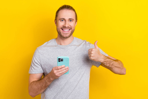 Photo of cheerful glad satisfied man wear stylish clothes arm thumb up quality gadget iphone isolated on yellow color background. - Foto, afbeelding