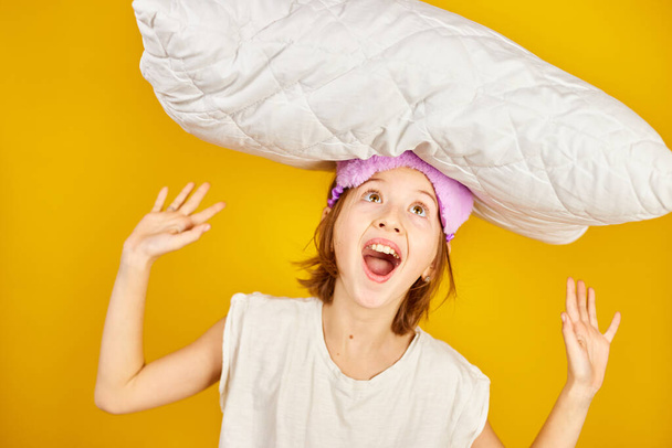 Funny teenage girl in white pyjamas with a violet sleeping mask hold pillow on the head, having fun, make faces in the studio on a yellow background - Фото, изображение