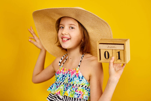 Lapsi teini tyttö uimapuku ja olki hattu pidä kädessä puinen kalenteri 01 Kesäkuu studiossa keltaisella pohjalla, kesän alku, kesätunnelma, Maailman lasten päivä - Valokuva, kuva