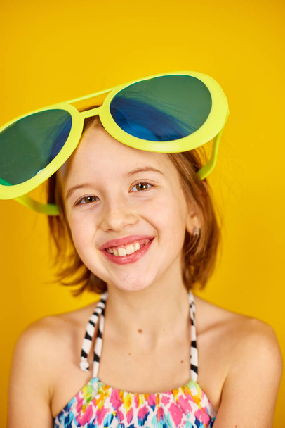 Smilling Child teenager girl in swimsuit and big sunglasses posing, having fun, make faces in studio on yellow background, summer mood, - Zdjęcie, obraz