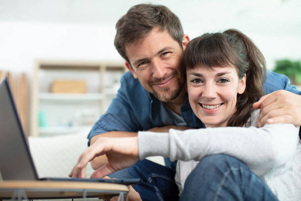 optimistic hugging young loving couple near sofa using laptop computer - Foto, afbeelding