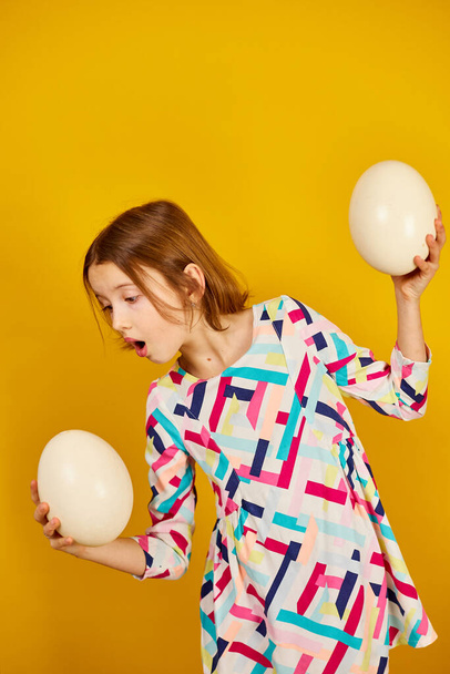 Happy playful teenager girl holding Ostrich eggs on a bright yellow studio background, celebrating Easter in style. - Фото, изображение