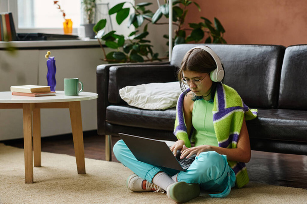Дівчина-підліток в навушниках сидить на підлозі у вітальні і вивчає онлайн на ноутбуці
 - Фото, зображення