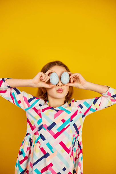 Happy playful teenager girl holding painted Easter eggs covering eyes, on a bright yellow studio background, celebrating Easter in style. - Foto, Imagen