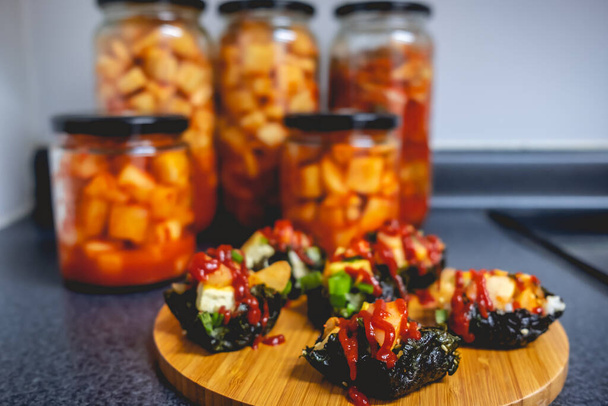 Wooden tray with korean-inspired snacks of kimchi, tofu, green onion, gochujang (hot pepper paste) and nori, and glass jars with kimchi in the background - Photo, Image
