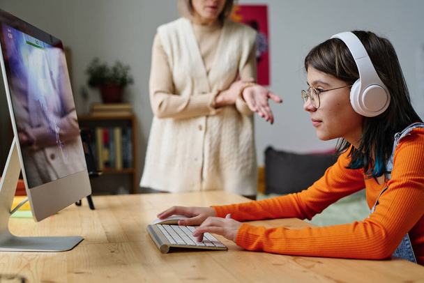 若いです女の子でワイヤレスヘッドフォンでテーブルの前に座っていますコンピュータモニタと彼女の母親とコンピュータゲームをプレイ彼女に発言をします - 写真・画像