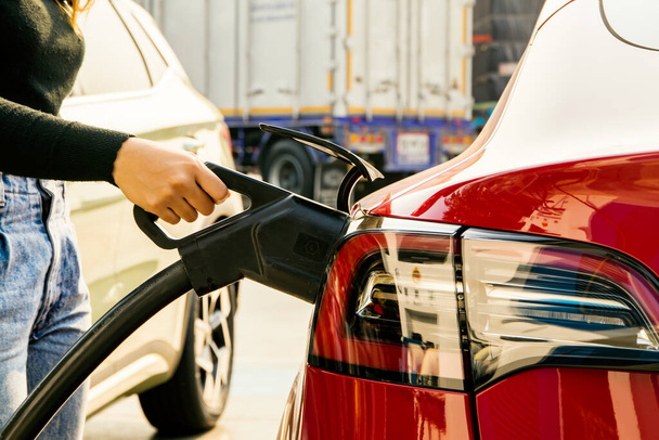 Fiatal női EV vagy elektromos autó vezető kezét tartja DC tápcsatlakozó az ő piros elektromos autó akkumulátor aljzat benzinkút: Tiszta energia jármű Alternatív energia egyre népszerűbb koncepció. - Fotó, kép