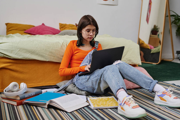 Школярка сидить на підлозі серед книг у своїй кімнаті з ноутбуком на колінах і навчається онлайн
 - Фото, зображення