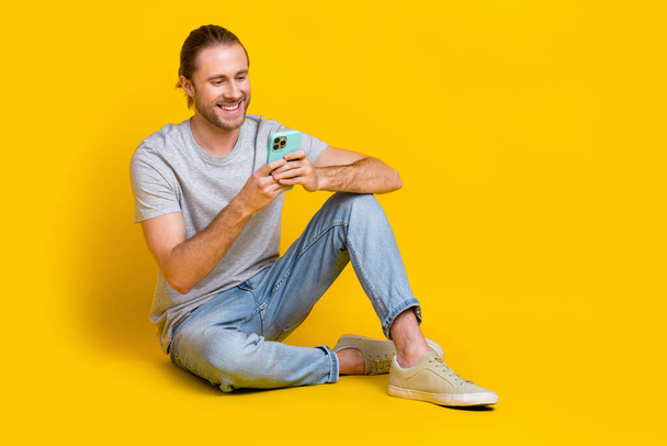 Full length photo of good mood optimistic guy dressed light t-shirt jeans look at smartphone chatting isolated on yellow color background. - Zdjęcie, obraz