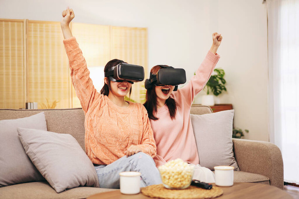 Jovem casal de lésbicas vestindo suéter e óculos VR para levantar o braço com o rosto animado enquanto assiste experiência realidade virtual com desfrutar e passar tempo juntos na sala de estar em casa. - Foto, Imagem