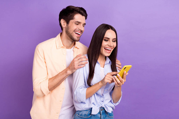 Photo of overjoyed funky couple husband hugs his wife together watching smartphone youtube trend video joke isolated on violet color background. - Fotó, kép