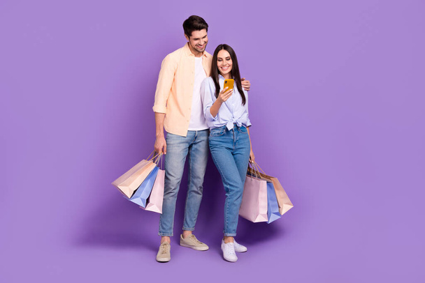 Full length portrait of two attractive idyllic partners embrace hold bags smart phone online order isolated on purple color background. - Fotoğraf, Görsel