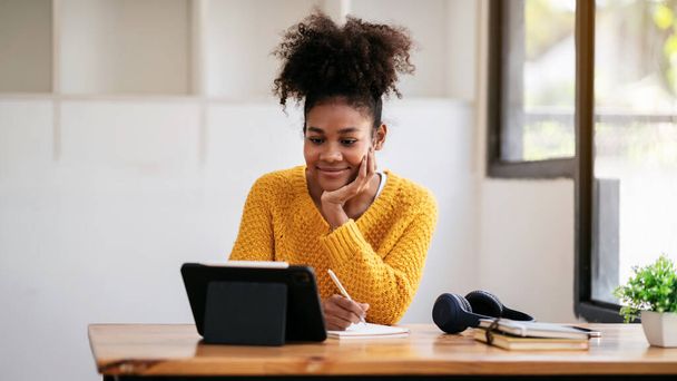 Afrikai amerikai diák nő pulóverben nézi e-learning tanulni leckét online osztály táblagépen, és jegyzetel, miközben ül az asztalon tanulni tudás és oktatás az egyetemen. - Fotó, kép