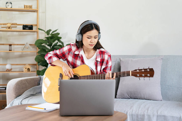 Genç kadın gitar çalma ve kulaklık takıp dizüstü bilgisayarla müzik aleti çalarken oturma odasındaki kanepede rahat bir şekilde oturmaya çalışıyor.. - Fotoğraf, Görsel