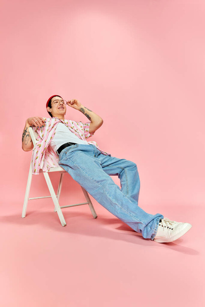 Disattento, divertito. Ragazzo alla moda sorridente e appoggiato indietro e in posa mentre seduto sulla sedia su sfondo rosa. Concetto di godimento della vita, emozioni umane, moda, gioventù, vita studentesca - Foto, immagini