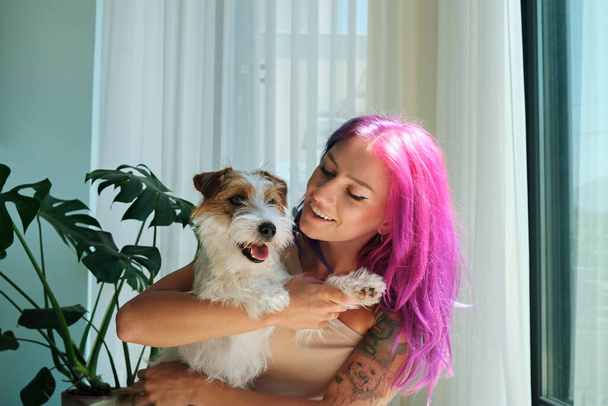 Young beautiful woman with pink hair hugging with her wire haired jack russell terrier at home. Broken coated pup in arms of smiling female. Close up, copy space, background. - Zdjęcie, obraz
