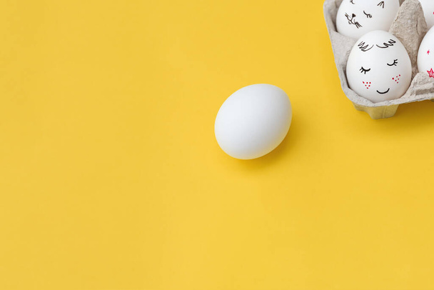 Carte de Pâques. Un œuf blanc sur fond jaune vif. Œufs peints avec des sourires dans le plateau. Vue supérieure avec espace de copie. Joyeuses fêtes. - Photo, image