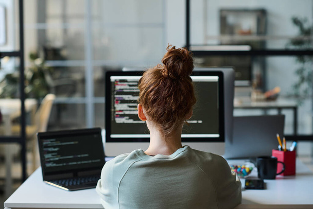 Vue arrière d'une jeune programmeuse assise sur son lieu de travail devant un écran d'ordinateur et tapant des codes pour un programme - Photo, image