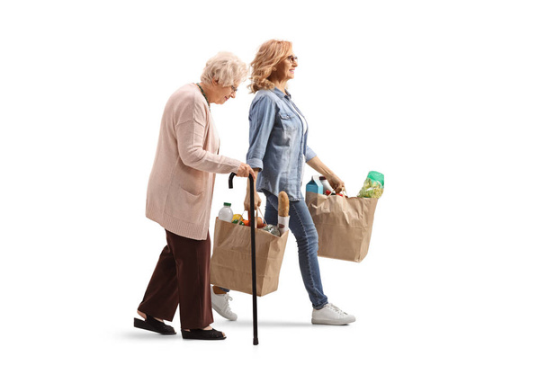 Full length profile shot μιας ηλικιωμένης γυναίκας που περπατάει με μπαστούνι και μια νεότερη γυναίκα που κουβαλάει τσάντες παντοπωλείου απομονωμένες σε λευκό φόντο - Φωτογραφία, εικόνα