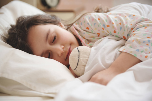 Κοντινό πλάνο πορτραίτο ενός πεντάχρονου Καυκάσιου αξιαγάπητου μικρού κοριτσιού με πιτζάμες με πολύχρωμες τελείες, αγκαλιάζει απαλά το βελούδινο πρόβατο παιχνίδι της, ενώ κοιμάται στο κρεβάτι σε ένα ελαφρύ υπνοδωμάτιο εσωτερικό. Παιδική ηλικία - Φωτογραφία, εικόνα