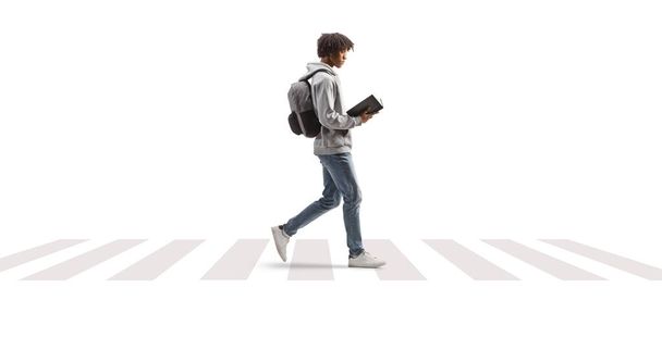 Full length profile shot of a young african american man walking and reading a book at a pedestrian crossing isolated on white background - Foto, afbeelding