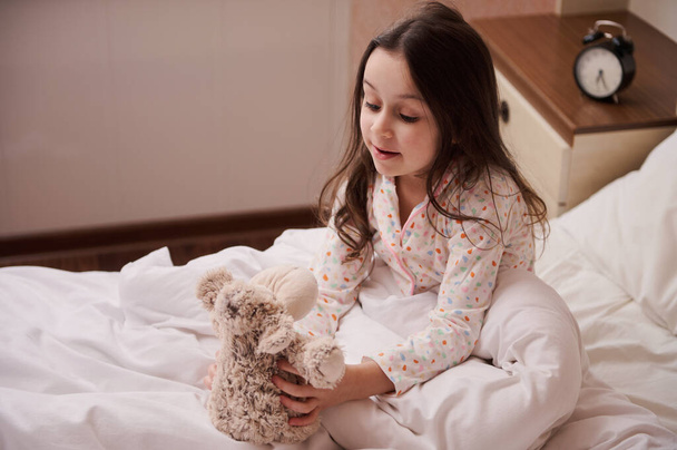 Adorable Caucasian little child girl with beautiful long hair, dressed in stylish pajamas, sitting on the bed in her bedroom and playing with her plush toy sheep, before going to bed. Kids. Childhood - Foto, afbeelding