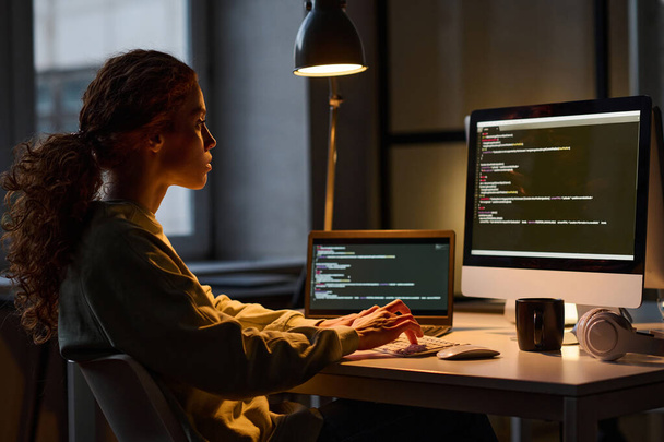 Молода жінка сидить на робочому місці і працює на комп'ютері з кодами безпеки в темному офісі
 - Фото, зображення
