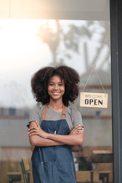 Erfolgreiche Afrikanerin in Schürze vor der Tür eines Cafés. Glücklicher Kleinunternehmer, der Tablet hält und arbeitet. Lächelndes Porträt eines KMU-Unternehmers, der mit Kopierraum dasteht. Hoch - Foto, Bild