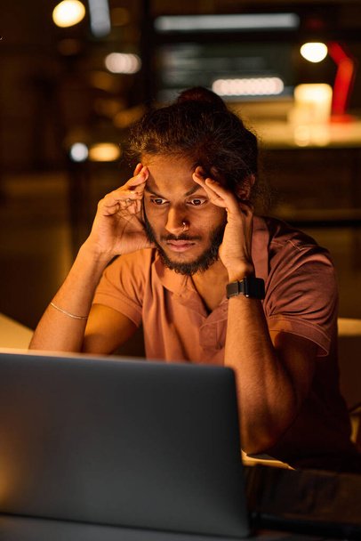 Imagen vertical del joven programador que se ve muy cansado sentado en su lugar de trabajo, que trabaja sobre el programa de ordenador en la oficina oscura - Foto, imagen