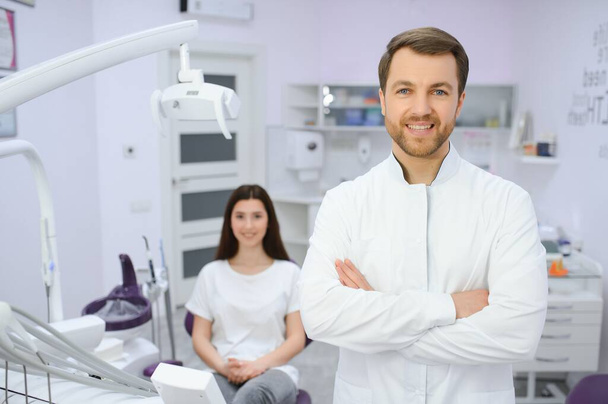 Odontólogo masculino en una habitación con equipo médico y paciente en segundo plano - Foto, Imagen