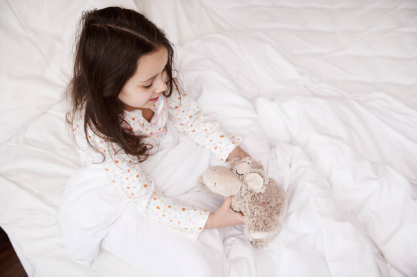 Vista dall'alto di una ragazza caucasica carina vestita in pigiama elegante con punti colorati, giocando con le sue pecore peluche giocattolo prima di andare a letto. Copia spazio pubblicitario - Foto, immagini