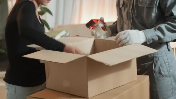 Przycięte ujęcie kobiety pakującej książki do pudełka kartonowego i przenoszącej pracownika serwisu uszczelniającego je taśmą samoprzylepną podczas przygotowywania się do opuszczenia domu - Materiał filmowy, wideo