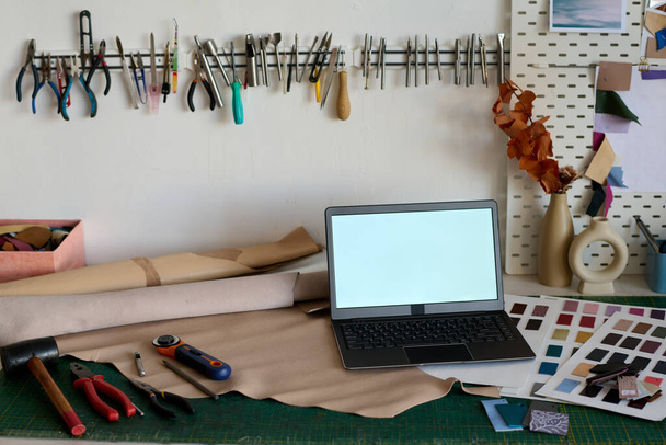 Laptop com tela em branco em pé no local de trabalho do curtidor com amostras de couro, ferramentas manuais, papel laminado para esboços e outras coisas - Foto, Imagem