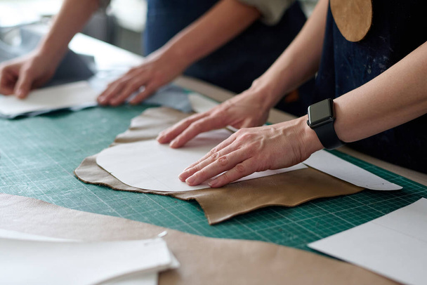 Primer plano de las manos de dos sastres o curtidores femeninos que ponen patrones de costura en piezas de cuero o gamuza mientras crean un nuevo atuendo en el taller - Foto, imagen