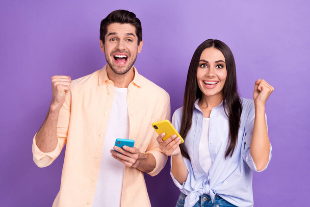 Photo of crazy overjoyed two people fists up wear casual stylish shirts celebrate good news read smartphone bet isolated on violet color background. - Фото, зображення