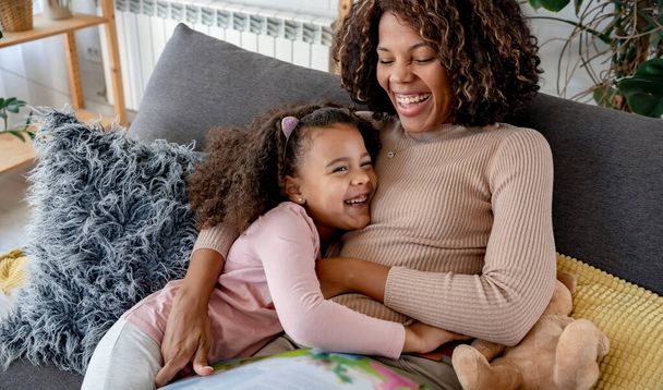 Αφροαμερικανή μητέρα και κόρη αγκαλιάζονται στο σπίτι στον καναπέ - Φωτογραφία, εικόνα