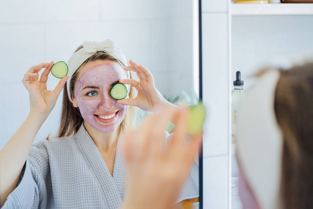 A young smiling woman with pink clay facial mask holds cucumber slices making a refreshing eye mask in bathroom. Natural cosmetic procedures for skin care at home. Beauty self-care. Selective focus. - Foto, afbeelding