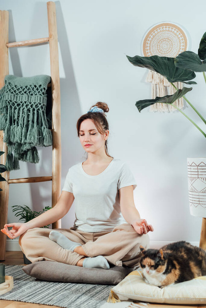 Young healthy woman practicing meditation at home, sitting in lotus position on floor cushion with relaxed cat pet in modern Scandi interior home. Meditating, relaxed, eyes closed. Vertical card - Foto, imagen
