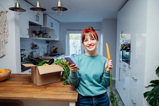 Lächelnde Frau mit frischer Karotte aus ihrer Lieferbox für gesunde Lebensmittel und ihrem Telefon mit Bestellung. Box mit Gemüse auf dem Küchentisch. Beginn eines gesunden Lebenskonzepts. Online-Essenslieferung nach Hause - Foto, Bild