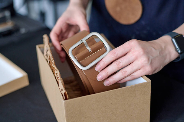 Ruce mladé fenky tanner uvedení nové ručně skládaný kožený pásek hnědé barvy do lepenkové krabice při balení pro klienta - Fotografie, Obrázek