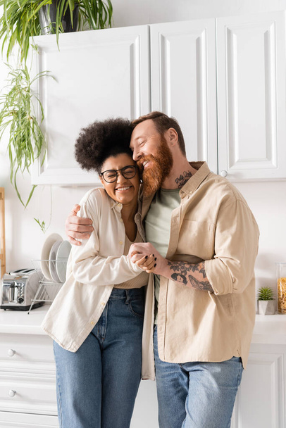 Smiling tattooed man hugging and holding hands with african american girlfriend in kitchen  - Foto, Bild